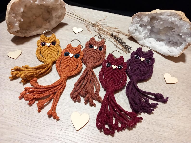 May include: A collection of handmade macrame owl keychains in various warm colors, including mustard yellow, orange, and burgundy. Each owl has a silver keyring, black eyes, and a small wooden heart. The keychains are displayed on a light wooden surface.