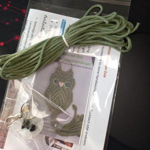 May include: A macrame owl keychain kit with green cord, a keychain, and instructions. The owl has a heart-shaped nose and glitter accents. The kit includes the text "kramee Box Schl&uuml;sselanh&auml;nger".