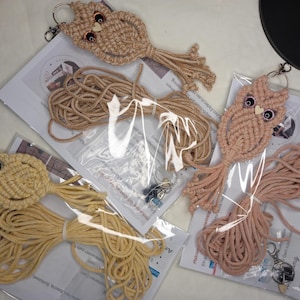 Könnte beinhalten: Makramee-Eulen-Schlüsselanhänger in Beige, Hellbraun und Pfirsich. Jede Eule hat einen Schlüsselring und ist mit Makramee-Kordel und Anleitungen verpackt. Die Eulen haben Knopfaugen und eine herzförmige Holznase. Die Schlüsselanhänger liegen auf bedrucktem Papier.