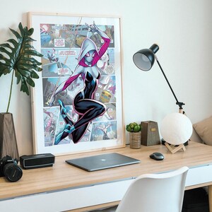 May include: A framed comic book poster featuring a female superhero in a white and black suit with pink accents. She is swinging from a web and has a white mask with a spider design. The poster is hanging on a white wall above a desk with a laptop, a lamp, and a white chair.