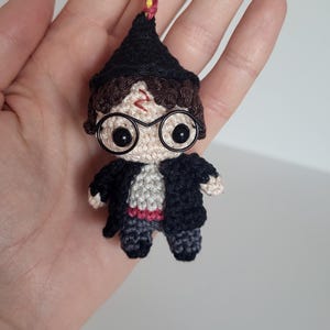Könnte beinhalten: Häkel-Schlüsselanhänger Harry Potter mit schwarzem Umhang, runden Brillen und einem braunen Hut. Der Schlüsselanhänger ist an einer rot-gelben Schnur befestigt.