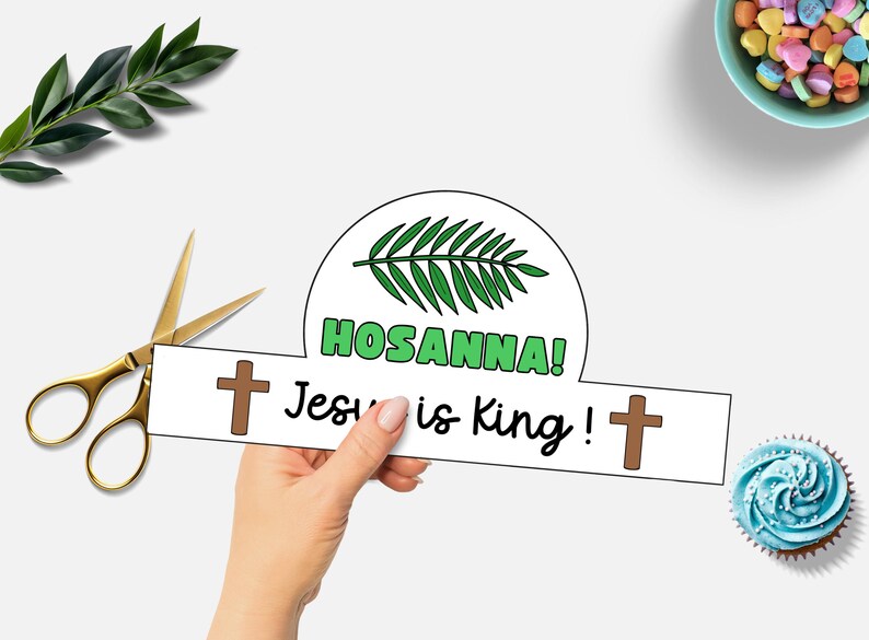 May include: A paper crown with the words "HOSANNA! Jesus is King!" and two brown crosses. The crown has a green palm branch design. Gold scissors and a sprig of greenery are next to the crown. A cupcake and a bowl of candy hearts are also in the image.
