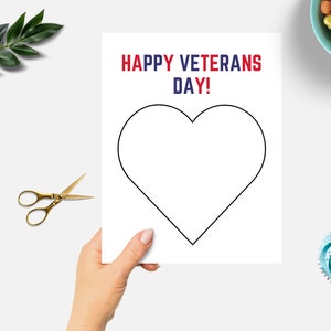 K&ouml;nnte beinhalten: Eine wei&szlig;e Karte mit einer schwarzen Herzumrandung, gehalten von einer Hand. Der Text "HAPPY VETERANS DAY!" ist in Rot und Blau gedruckt. Goldene Schere und ein Zweig gr&uuml;ner Bl&auml;tter befinden sich links. Eine Schale mit S&uuml;&szlig;igkeitenherzen und ein Cupcake befinden sich rechts.