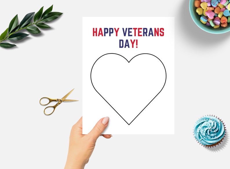 K&ouml;nnte beinhalten: Eine wei&szlig;e Karte mit einer schwarzen Herzumrandung, gehalten von einer Hand. Der Text "HAPPY VETERANS DAY!" ist in Rot und Blau gedruckt. Goldene Schere und ein Zweig gr&uuml;ner Bl&auml;tter befinden sich links. Eine Schale mit S&uuml;&szlig;igkeitenherzen und ein Cupcake befinden sich rechts.