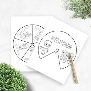 May include: Two white paper cutouts with black line drawings. One cutout is a circle divided into sections with illustrations and text. The other cutout has the name "STEPHEN" and a drawing of a man. A gold pen rests on the paper.