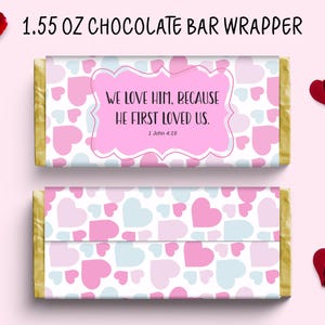 Op de afbeelding: Chocoladereepverpakking met een patroon van roze en lichtblauwe harten. De verpakking toont de tekst "WE LOVE HIM, BECAUSE HE FIRST LOVED US." en "1 John 4:19" in een roze wolk-tekstvak. De verpakking is gelabeld "1.55 OZ CHOCOLATE BAR WRAPPER".