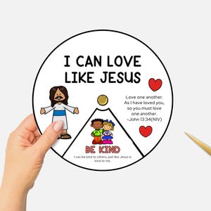 May include: A circular graphic with the text "I CAN LOVE LIKE JESUS" and "BE KIND". The image includes illustrations of Jesus, children, and hearts. A hand holds the graphic, with gold scissors in the background.