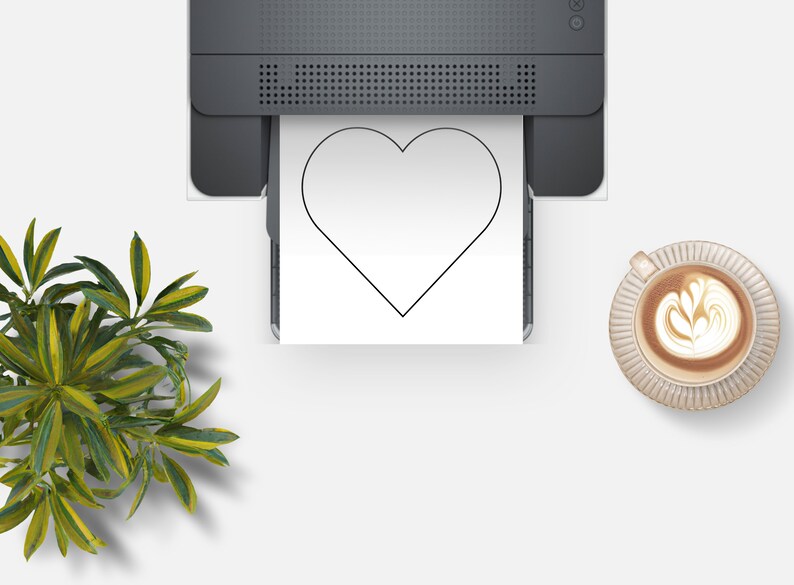 K&ouml;nnte beinhalten: Draufsicht auf einen Drucker, der eine gro&szlig;e schwarze Herzform auf wei&szlig;em Papier druckt. Rechts eine Tasse Kaffee mit Latte Art und links eine gr&uuml;ne Pflanze.