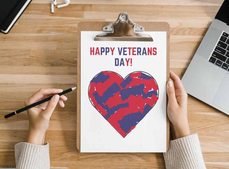 K&ouml;nnte beinhalten: Ein Klemmbrett mit einem wei&szlig;en Blatt Papier mit dem Text "HAPPY VETERANS DAY!" in Rot und einem Herzdesign, das mit roten und blauen Pinselstrichen gef&uuml;llt ist. Eine Hand h&auml;lt einen Bleistift, und ein Laptop ist sichtbar.