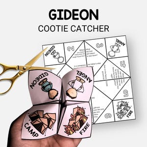 May include: A folded paper fortune teller with the word "GIDEON" and "COOTIE CATCHER" printed on it. The fortune teller is pink and white with illustrations and text. A pair of gold scissors is next to the fortune teller.