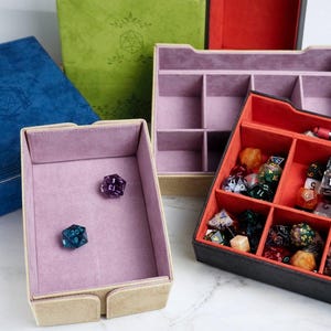 May include: A collection of rectangular storage boxes for gaming dice, featuring various exterior colors including blue, green, and black. The interiors are lined with soft fabric in shades of lavender and red, with divided compartments for organizing dice.
