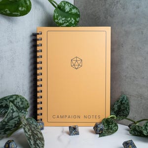 May include: A yellow spiral bound notebook with the text "Campaign Notes" on the front cover. The notebook has a black geometric design on the front cover. The notebook is surrounded by black and grey dice.