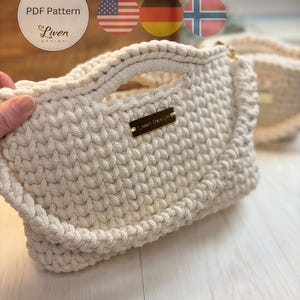 May include: A cream-colored crocheted handbag featuring a thick, textured stitch pattern, integrated handles, and a rectangular gold-toned metal plaque engraved with the text Liven Design. A circular label displays the text PDF Pattern Liven Design.