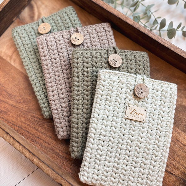 May include: Four crocheted phone cases in different colors, including green, beige, and cream. Each case has a wooden button and a wooden tag with the word "Liven" on it.