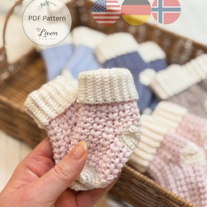 Op de afbeelding: Handgemaakte babysokjes in zachtroze en crème. De sokjes zijn gehaakt met een textuursteek en hebben een omgeslagen boord. Andere sokjes zijn zichtbaar in een rieten mand. De tekst "PDF Pattern Liven Design" is zichtbaar.