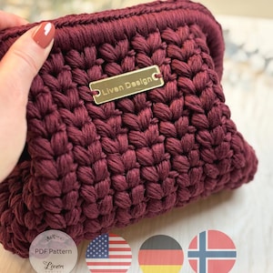 May include: A burgundy crochet clutch bag with a textured stitch pattern. A gold-coloured rectangular tag is attached, bearing the inscription "Liven Design". The clutch is held by a hand with painted nails. The image includes a "PDF Pattern" logo and flags.