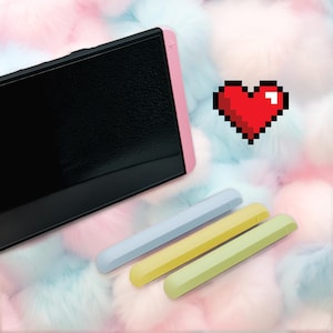 Pode incluir: Um tablet de escrita digital preto com uma borda rosa e três canetas de cores pastel. Um gráfico de coração vermelho pixelado também está presente. O tablet tem uma superfície de escrita texturizada.