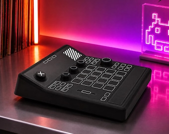 May include: A black audio mixing console featuring a grid of square buttons, several circular control knobs, a large cross-marked button on the left, and a rectangular slider. The device has a slanted top surface with white outlines for each control.