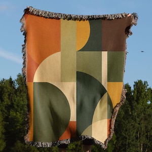 May include: A woven throw blanket featuring a geometric pattern of overlapping circles and rectangles in shades of burnt orange, mustard yellow, olive green, dark brown, and cream. The blanket has a fringed edge along the top and bottom sides.
