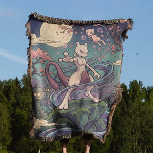 May include: A woven throw blanket featuring a fantasy landscape with a full moon, cherry blossoms, and two Pokémon characters. The blanket has a blue, purple, and pink color scheme with a fringed edge.