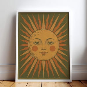 May include: A framed rectangular wall art print featuring a central sun and moon face illustration. The design displays a golden yellow celestial face with rosy cheeks against a dark olive green background, surrounded by radiating orange sun rays.