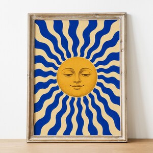 May include: A framed wall art print featuring a central yellow sun with a human face, surrounded by alternating wavy blue and cream-colored rays radiating outward. The artwork is enclosed in a light-toned wooden frame with a rectangular shape.
