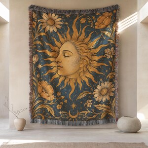 May include: A woven throw blanket featuring a dark blue background with a large, central golden sun face in profile. The design includes intricate floral motifs, stars, and a crescent moon at the bottom, finished with a decorative fringed edge.
