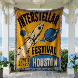 May include: A yellow woven throw blanket with fringed edges featuring a black graphic of a rocket ship, a blue planet with rings, and a smaller blue planet. The text reads INTERSTELLAR FESTIVAL HOUSTON and the dates JUNE 3 25-27.