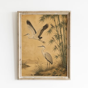 May include: A framed wall art print featuring a tan background with two grey and white herons. One heron is in flight while the other stands near a riverbank. Green bamboo stalks and small birds are visible on the right side of the illustration.