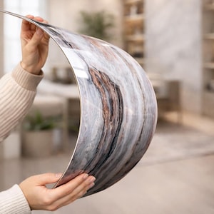 Può includere: Un foglio sottile e flessibile con finitura lucida e motivo marmorizzato nei toni del bianco, grigio, nero e rame. Il materiale è curvato a mezzaluna, mostrando una superficie liscia e riflettente con venature intricate e fluide.
