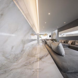 May include: Interior shot of a modern lounge area with a long, gray sofa and marble flooring. The wall is white with marble veining, and the ceiling has recessed lighting. Decorative pillows line the sofa.
