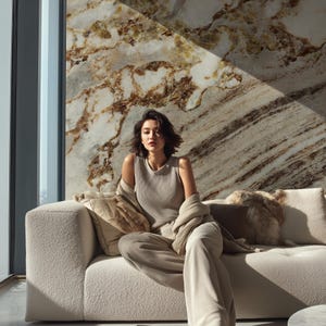 Op de afbeelding: Een vrouw in een beige mouwloze top en bijpassende broek zit op een crèmekleurige bank. Een lichtbruin vest is over haar schouders gedrapeerd. De achtergrond is een marmeren muur met bruine en beige aderen en een groot raam.