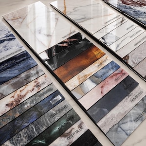 May include: A collection of rectangular stone tile samples arranged in a grid, featuring various marble and onyx patterns in colors including white, blue, pink, black, and gold. Each tile is labeled with names like Pink Powder, Onyx Gold, and Travertino.