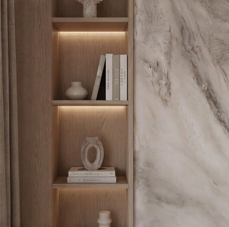 May include: A wooden bookshelf with built-in lighting displays decorative items. The shelves hold books, vases, and a carved decorative piece. The background features a marble wall.