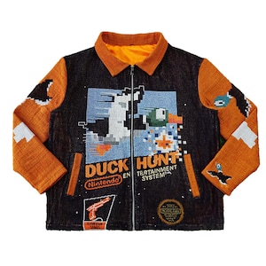 Pode incluir: Uma jaqueta preta e laranja com um design pixelado de um pato e uma arma. A jaqueta tem um fecho de correr e o texto "Duck Hunt Nintendo Entertainment System" na frente.