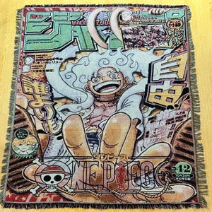 May include: A woven throw blanket with a vibrant manga-style illustration. The design features a central character with white hair and a wide smile, surrounded by Japanese text and the words "ONE PIECE". The blanket has a fringed edge.