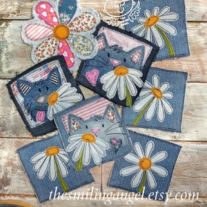 May include: A collection of denim fabric patches featuring embroidered designs of daisies and cats with pink hearts. The patches include a large multi-patterned flower shape and several square denim pieces with frayed edges and stitched floral motifs.