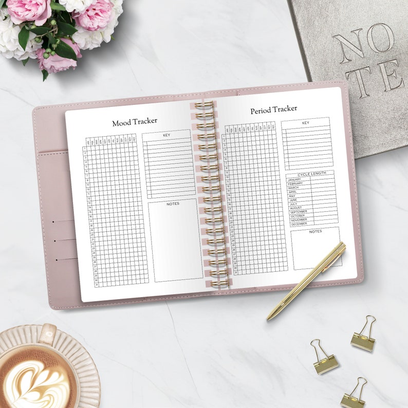 Puede incluir: Diario rosa abierto con encuadernaci&oacute;n en espiral dorada, con p&aacute;ginas de "Mood Tracker" y "Period Tracker". Un bol&iacute;grafo dorado y clips est&aacute;n sobre la superficie de m&aacute;rmol. Una taza de caf&eacute; y un ramo de flores rosas y blancas tambi&eacute;n est&aacute;n presentes.