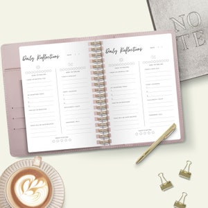 K&ouml;nnte beinhalten: Ein rosafarbenes, ledergebundenes Tagebuch mit goldener Spiralbindung. Das Tagebuch tr&auml;gt den Schriftzug "Daily Reflections" auf dem Einband. Die Seiten sind mit Aufforderungen zur t&auml;glichen Reflexion und Dankbarkeit gef&uuml;llt.