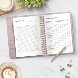 May include: A pink leather-bound journal with a gold spiral binding. The left page has a gridded sleep tracker with the heading "Sleep Tracker". The right page has a gridded "Improving Sleep" tracker with headings for "Time I went to bed", "Time I turned off light", "Time it took to fall asleep", "Number of times I woke up in the night", "Number of times I got up to the toilet", "Time I woke up", "Total time asleep", "Total time I was restless in the night", "Total time I spent awake", "Units of alcohol", "Sleeping pills", "Quality of sleep - marks out of 10", "Tiredness - marks out of 10".