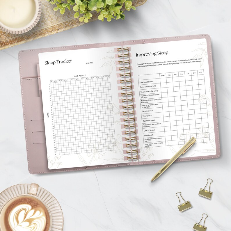 May include: A pink leather-bound journal with a gold spiral binding. The left page has a gridded sleep tracker with the heading "Sleep Tracker". The right page has a gridded "Improving Sleep" tracker with headings for "Time I went to bed", "Time I turned off light", "Time it took to fall asleep", "Number of times I woke up in the night", "Number of times I got up to the toilet", "Time I woke up", "Total time asleep", "Total time I was restless in the night", "Total time I spent awake", "Units of alcohol", "Sleeping pills", "Quality of sleep - marks out of 10", "Tiredness - marks out of 10".