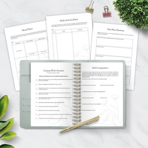 May include: A green and white journal with gold spiral binding. The journal has pages for daily activities, mood tracking, coping with anxiety, and self-compassion exercises. The pages are filled with prompts and checklists.