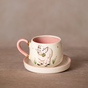 Könnte beinhalten: Ein Keramik-Tassen- und Untertassen-Set. Die Tasse ist cremefarben mit rosa Innenraum und Henkel, akzentuiert mit goldenen Verzierungen. Ein rosa Schwein mit einem vierblättrigen Kleeblatt befindet sich auf der Tasse. Die Untertasse ist cremefarben mit rosa Rand.