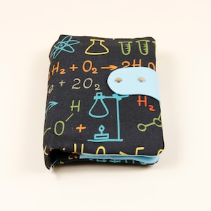 May include: A black fabric wallet with a science theme. The wallet has a blue flap with two silver snaps. The fabric has a pattern of colorful chemical formulas, beakers, and test tubes.