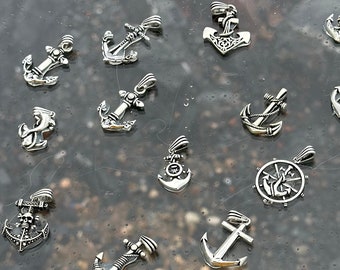 May include: A collection of silver-toned anchor charms, some with intricate details, displayed on a glass surface. The charms are various sizes and styles, including a compass and a skull and crossbones.