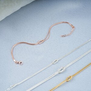 Pode incluir: Tr&ecirc;s delicados braceletes de corrente de ouro, prata e ouro rosa com um design de n&oacute;. Os braceletes est&atilde;o dispostos sobre um fundo de tecido azul claro.