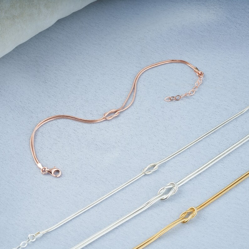 Pode incluir: Tr&ecirc;s delicados braceletes de corrente de ouro, prata e ouro rosa com um design de n&oacute;. Os braceletes est&atilde;o dispostos sobre um fundo de tecido azul claro.