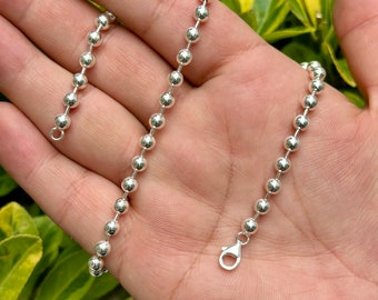 May include: A silver ball chain necklace with a lobster clasp, held in a hand. The necklace is composed of small, round, shiny beads. The background is a blurred view of green and yellow foliage.
