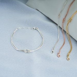 Pode incluir: Tr&ecirc;s pulseiras de corrente prateadas, de ouro rosa e de ouro com um design de n&oacute;. As pulseiras est&atilde;o sobre um fundo de tecido azul claro.