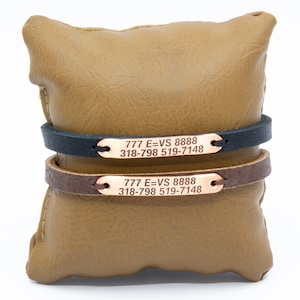 Puede incluir: Dos pulseras de cuero con una placa de metal de oro rosa grabada con el texto "777 E=VS 8888 318-798 519-7148". Una pulsera es de cuero negro y la otra de cuero marrón.
